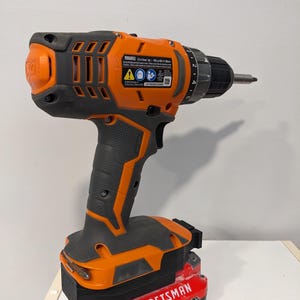 May include: An orange and black cordless drill with a red 20V Craftsman battery attached. The drill has a black handle and a silver drill bit. The drill has a speed setting dial and a trigger.