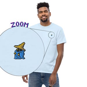 May include: Light blue t-shirt featuring a pixelated wizard design. The design is in blue and yellow, with a zoomed-in view of the graphic. The shirt is a classic crew neck style.