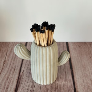 Puede incluir: Un portafósforos en forma de cactus gris claro lleno de fósforos de madera. El portafósforos tiene una textura acanalada y dos brazos laterales. Los fósforos tienen puntas negras y están colocados en posición vertical en el portafósforos. El soporte está sobre una superficie de madera.