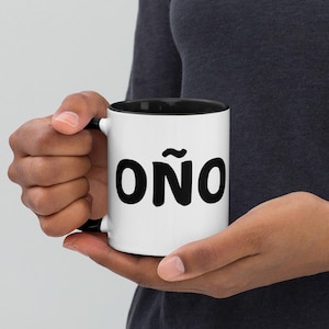 May include: White ceramic mug with a black rim and handle. The mug has a black graphic of the word "OÑO" with a tilde over the "O".