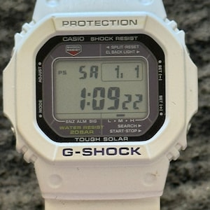 May include: A white G-Shock watch with a black digital display showing the time, date, and day of the week. The watch has a black bezel and a white strap. The watch face also displays the words "Protection", "Shock Resist", "Water Resist 20BAR", "Tough Solar", and "G-Shock".