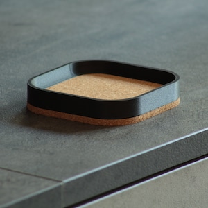 Puede incluir: Un posavasos cuadrado negro con un inserto de corcho. El posavasos está hecho de un material plástico duro y tiene un acabado liso.