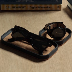 Puede incluir: Dos bandejas negras forradas de corcho con bordes redondeados. Una bandeja contiene un par de gafas de sol negras, la otra contiene un reloj de pulsera negro con una correa de cuero marrón.