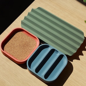 May include: Three different coloured trays made of a textured material. The trays are in shades of green, blue, and pink. The green tray has a rippled texture. The blue tray has three indented sections. The pink tray has a cork surface.