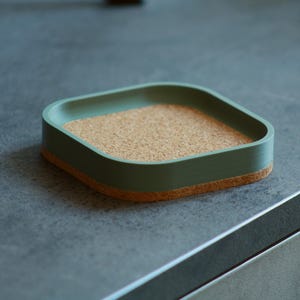 Może przedstawiać: Kwadratowa, zielona podkładka pod kubek z wkładką korkową. Podkładka wykonana jest z gładkiego, twardego materiału i ma lekko zaokrąglone krawędzie.