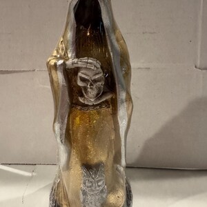May include: A gold-colored figurine of a hooded figure, with silver accents on the hood and edges. The figure holds a skull and crossbones, with an owl at the base. The statue is wrapped in clear plastic.