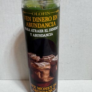 May include: A tall, cylindrical glass candle with a green wax interior and a clear lid. The label features text in Spanish and English, including "See Money in Abundance" and images of money bags and stacks of bills. The candle is designed to attract money and abundance.