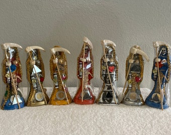May include: Set of seven colorful skeletal figures, each holding a scythe and adorned with gold accents and various trinkets. The figures are standing on a white surface.