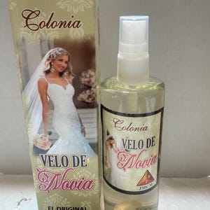 Może przedstawiać: Przezroczysta szklana butelka wody kolońskiej i pasujące pudełko. Na butelce i pudełku widnieją napisy "Colonia Velo De Novia" oraz wizerunek panny młodej w sukni ślubnej. Butelka zawiera 100 ml naturalnego sprayu.