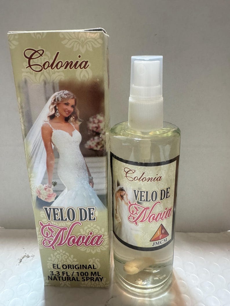 Może przedstawiać: Przezroczysta szklana butelka wody kolońskiej i pasujące pudełko. Na butelce i pudełku widnieją napisy "Colonia Velo De Novia" oraz wizerunek panny młodej w sukni ślubnej. Butelka zawiera 100 ml naturalnego sprayu.