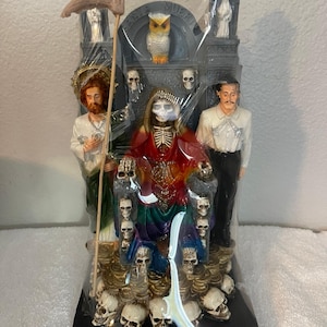 May include: A detailed figurine featuring a central skeletal figure in a red and rainbow robe, surrounded by skulls and other figures. The statue includes an owl and the text "SANTA MUERTE". The base measures 11 3/4 inches.