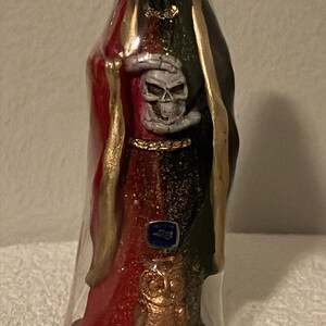 May include: A tall, decorative candle in a plastic wrap. The candle is shaped like a robed figure with a skull face, wearing a red and black robe with gold accents. The figure is holding a skull. The base is dark.
