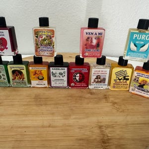 May include: A collection of eleven small rectangular glass bottles filled with various colored spiritual oils. Each bottle features a black cap and a unique label with text and imagery, including titles like Pusanga del Amor, Sandalo, Ven a Mi, Puro, 7 Monedas, Destrancadera, Gambling, Reversible, Leche de la Mujer Amada, Contra Maleficios, and Abrir Caminos.