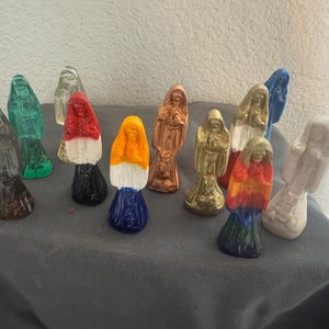 May include: A collection of small, hooded figurines in various colors, including green, red, gold, and blue. The figurines are arranged on a gray surface, with a neutral background.