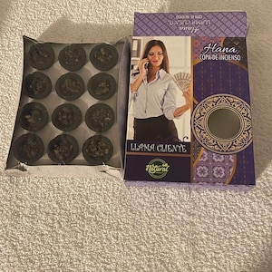 May include: A box of incense cups and a purple box with the text "Hana Copa de Incienso" and an image of a woman. The incense cups are dark brown and round. The box has a floral pattern and the word "Natural".