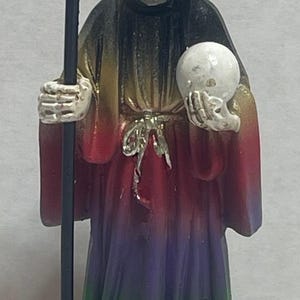Peut inclure: Figurine d'un squelette vêtu d'une robe, tenant un crâne et une faux. La robe est un dégradé de rouge, jaune, vert et violet. Une figurine de hibou est à la base, sur une pile de pièces d'or.