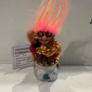 Op de afbeelding: Een grillig trollenbeeldje met roze en geel haar. De trol houdt een pot goud vast en zit op een zilveren pot met een groene klaver. Een kleine kaart met Spaanse tekst staat op de voorgrond.