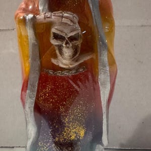 May include: A colorful, decorative candle shaped like a hooded figure. The candle features a skull and crossbones design on the front, with an owl at the base. The colors transition from red to yellow to blue, with silver accents.