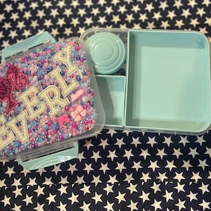 May include: A light blue lunch box with multiple compartments. The lid is decorated with beads in shades of pink, blue, and white, spelling out the name "EVERLY" with a pink graphic. The lunch box is open, revealing the interior compartments.