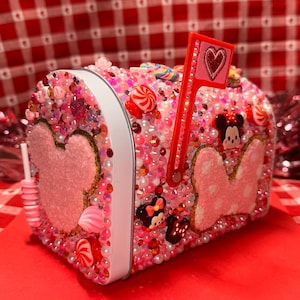 May include: A pink mailbox covered in beads, sequins, and candies. Features a Minnie Mouse design and a red flag with a heart. The mailbox is decorated with various candies and a lollipop.