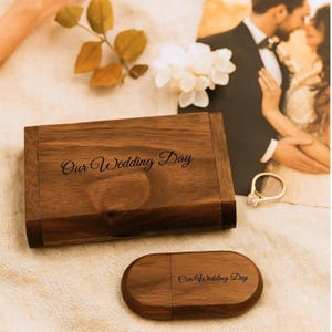 Puede incluir: Un conjunto de memoria USB y caja de madera con la inscripción "Our Wedding Day" grabada. La caja rectangular y la memoria USB ovalada son de madera oscura. Una foto de boda y un anillo se ven en el fondo.