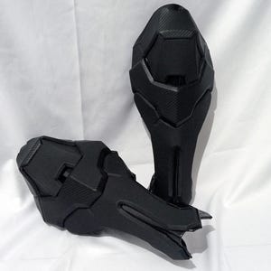 May include: Two black, futuristic-looking knee or shin guards. They have a textured, carbon fiber appearance and a segmented design. The guards are likely for protective gear or cosplay.