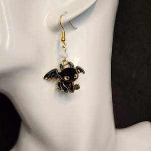 May include: A pair of gold-colored dangle earrings. Each earring features a black dragon charm with gold-colored accents and blue wings. A small, white bead is suspended between the charm and the gold-colored ear hook.