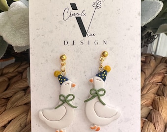 Puede incluir: Un par de pendientes caprichosos con figuras de gansos blancos con patas naranjas y un lazo verde. Cada ganso lleva un gorro de fiesta azul con lunares amarillos. Los pendientes cuelgan de postes dorados. La tarjeta dice "Clover & Vine DESIGN."