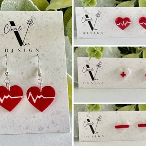 May include: Three sets of heart-shaped earrings. The first set is red with a white EKG line. The second set is white with a red cross. The third set is white with a red stripe. All earrings are on a white card with the text "Clover & Vine Design".