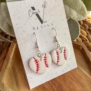 Könnte beinhalten: Herzförmige Ohrringe mit Baseball-Design. Die weißen Ohrringe haben rote Nahtdetails und sind an silberfarbenen Haken aufgehängt. Die Ohrringe werden auf einer weißen Karte mit dem Text "Clover & Vine DESIGN" präsentiert.