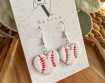 May include: Heart-shaped earrings with a baseball design. The white earrings have red stitching details, and are suspended from silver-colored hooks. The earrings are displayed on a white card with the text "Clover & Vine DESIGN".