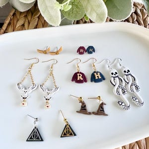 May include: A collection of handmade earrings on a white oval tray. The earrings feature various designs, including owls, sweaters with initials, wizard hats, and Deathly Hallows symbols. Colors include gold, black, white, brown, and burgundy.