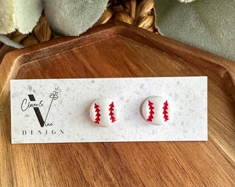 May include: A pair of white baseball stud earrings with red stitching. The earrings are presented on a white card, which features the text "Clover & Vine DESIGN". The card rests on a wooden tray with a visible wood grain.