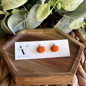 Puede incluir: Un par de pendientes de botón con forma de calabaza naranja con tallos marrones, presentados en una tarjeta blanca. La tarjeta tiene el texto "Clover & Vine Design". Los pendientes están sobre una bandeja de madera.