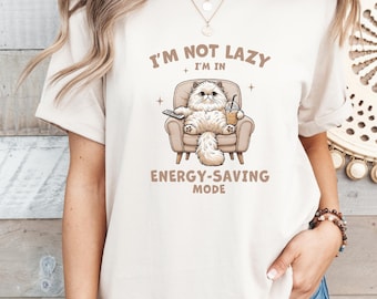 Peut inclure: T-shirt crème avec un chat de dessin animé se relaxant dans un fauteuil, tenant une télécommande et une boisson. Le texte dit "I'M NOT LAZY, I'M IN ENERGY-SAVING MODE". Le chat est duveteux et le fauteuil est marron clair.