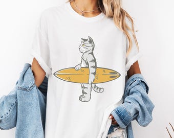 Puede incluir: Camiseta blanca con una ilustración de dibujos animados de un gato gris y blanco sosteniendo una tabla de surf amarilla. El gato está representado en un estilo simple y gráfico. La camiseta tiene un corte holgado y mangas cortas.