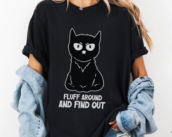 Pode incluir: T-shirt preta com uma ilustração branca de um gato zangado. O texto "FLUFF AROUND AND FIND OUT" está impresso abaixo da ilustração do gato. A camisa tem mangas curtas e um corte folgado.