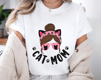 Peut inclure: T-shirt blanc avec un motif d'une femme aux cheveux bruns en chignon, portant un bandeau rose avec des oreilles de chat et des lunettes de soleil avec des silhouettes de chat. Les mots "CAT MOM" sont imprimés en noir en dessous.