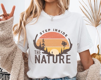 May include: White t-shirt with a nature-themed graphic. The design features a sunset scene with trees, a person, and the text "STEP INSIDE THE NATURE" in brown. The shirt is made of a soft material.