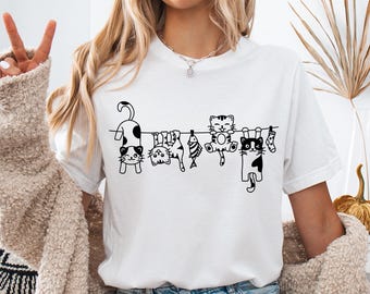 May include: White t-shirt featuring a black line drawing of several cats hanging from a clothesline. The cats have various patterns and are accompanied by a fish and a sock. The shirt is worn by a person.