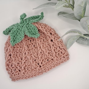 May include: A crocheted strawberry-shaped hat. The hat is a dusty rose colour, with a green crocheted leaf design at the top. The hat is likely for a baby or small child. The hat is made with soft yarn.