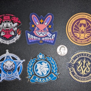 May include: A collection of eight colorful, stylized patches with various designs and text. The patches feature cartoon characters, logos, and text such as "GENTIE-HOUSE", "BELOBOG", and "H.S.O.S.6". A quarter is included for size reference.