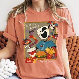 May include: Coral-colored t-shirt featuring a cartoon design with the text "Serve with a smile!" and "Try the sauce." The graphic includes a character holding a burger, fries, and a drink. The shirt has short sleeves.