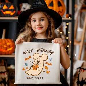 May include: A young girl in a black witch hat holds a white tote bag. The bag displays a cartoon ghost with a pumpkin bucket, and the text "WALT DISNEY EST. 1971". The design includes autumn leaves and Mickey Mouse-shaped ghosts.