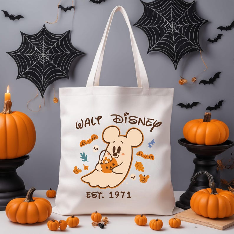 May include: A cream-coloured tote bag with a Halloween design. It features a cartoon ghost holding a pumpkin, with the text "Waly Disney EST. 1971". The background includes pumpkins, bats, and spiderwebs.