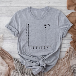 May include: A gray t-shirt with a black graph design. The graph shows a coordinate plane with the point (6,7) marked. The x-axis is labeled from 1 to 7, and the y-axis is labeled from 1 to 7.