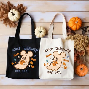 May include: Two tote bags, one black and one cream, with a Halloween design. The design features a ghost-like Mickey Mouse holding a pumpkin, the words "Walt Disney," and "Est. 1971." Pumpkins and autumn leaves surround the bags.
