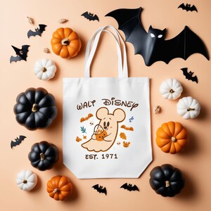 May include: White tote bag with a cartoon ghost Mickey Mouse holding a pumpkin bucket, with the text "Walt Disney EST. 1971". The image includes orange, black, and white pumpkins, and bat cutouts, perfect for Halloween.