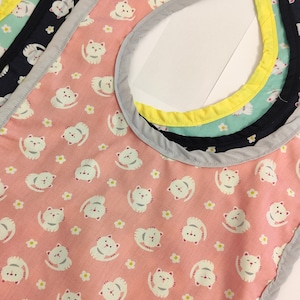 May include: A pink fabric bib with a white cat pattern and a multi-colored trim. The bib has a scalloped edge.