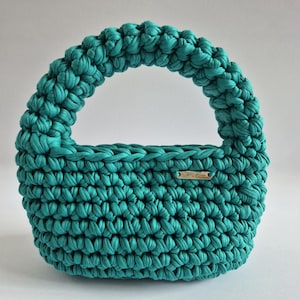 May include: A teal crochet handbag featuring a thick, rounded handle and a textured, knotted stitch pattern. A small, rectangular metallic tag with the engraved text "STPVA" is attached to the front of the bag, which has a wide, curved base.